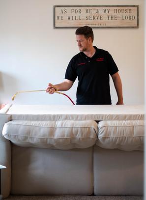 A professional technician performing a targeted pest control spray treatment behind a living room sofa.